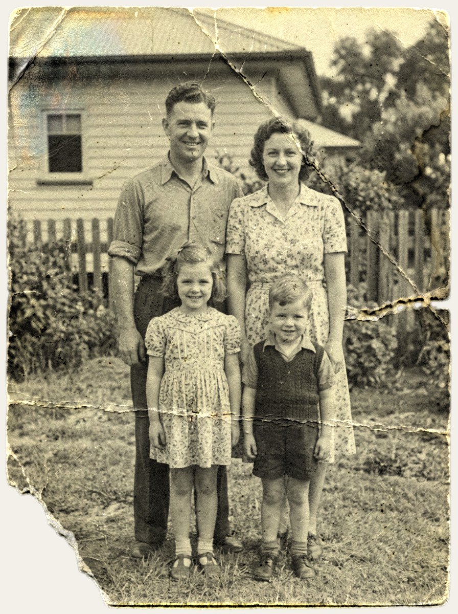Damaged 1940s black-and-white photograph of parents and two young children in a backyard, torn and creased with stains