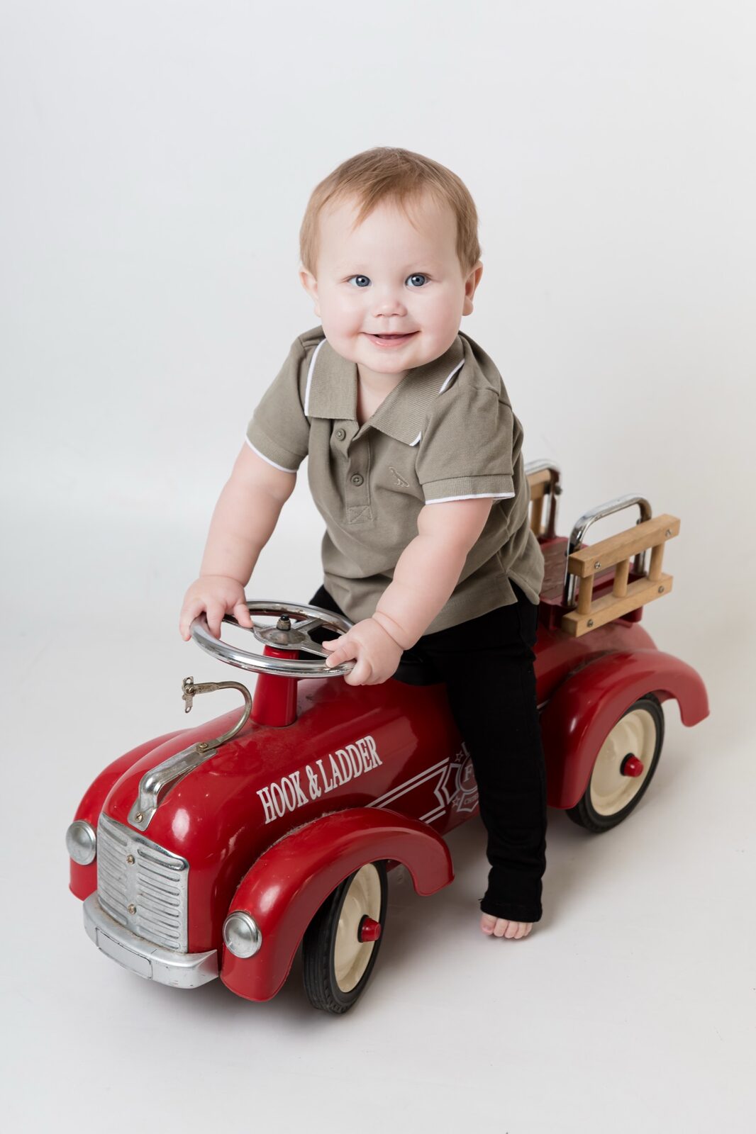 Smiling toddler in olive polo and black jeans sitting on a vintage red metal fire truck ride-on in a studio