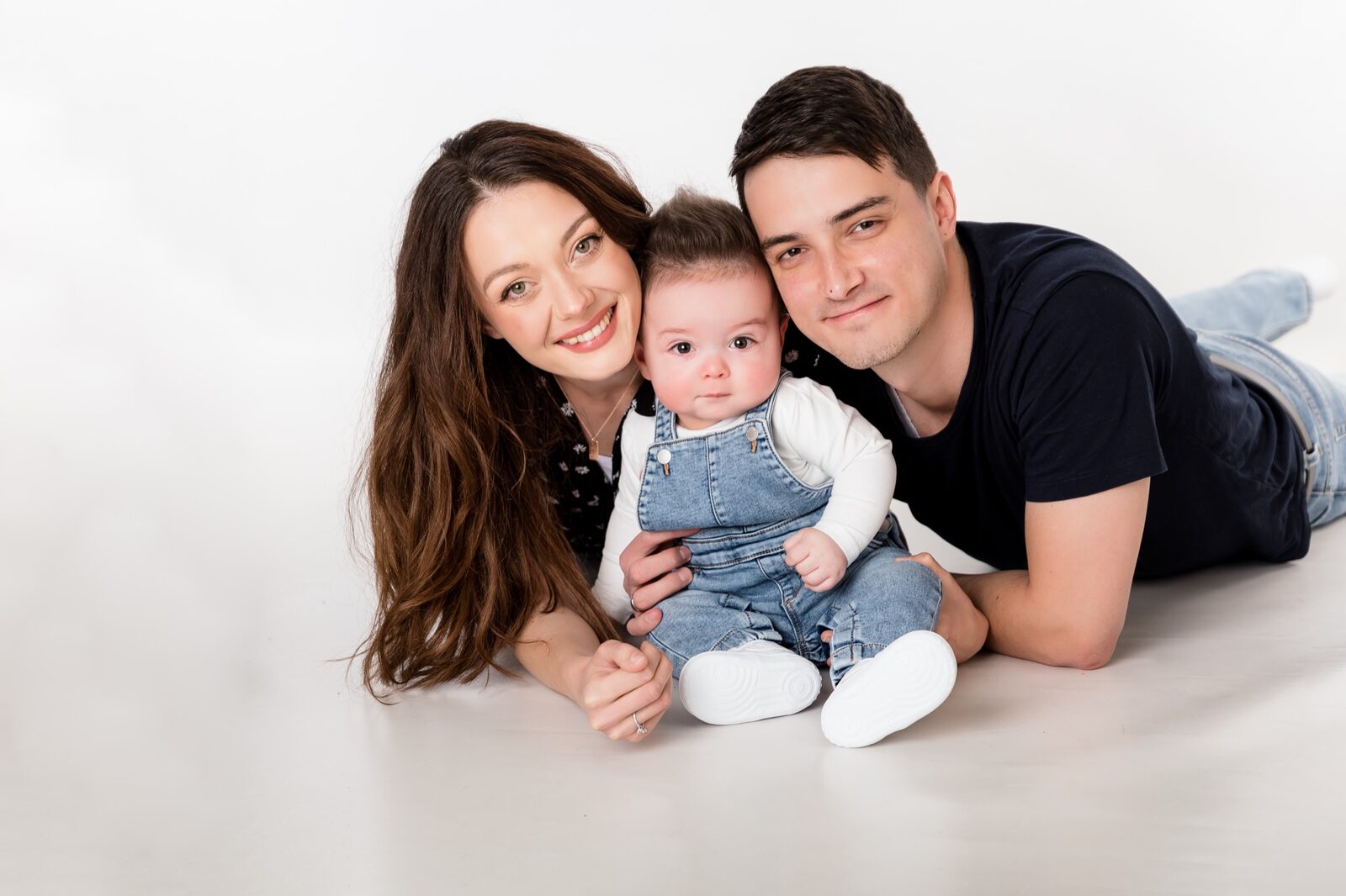 Mother and father lying on white studio floor cuddling their baby dressed in denim overalls and white shoes