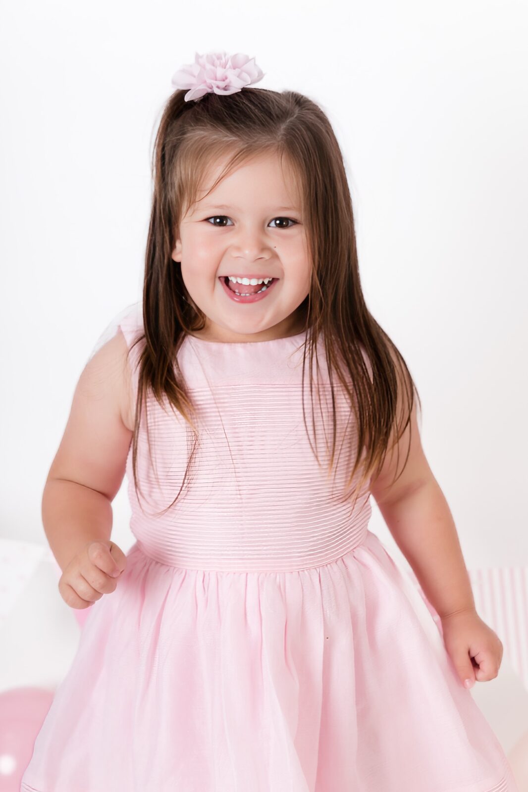 Smiling toddler girl with long brown hair and a pink flower hair clip, wearing a pink dress, photographed on a white studio…