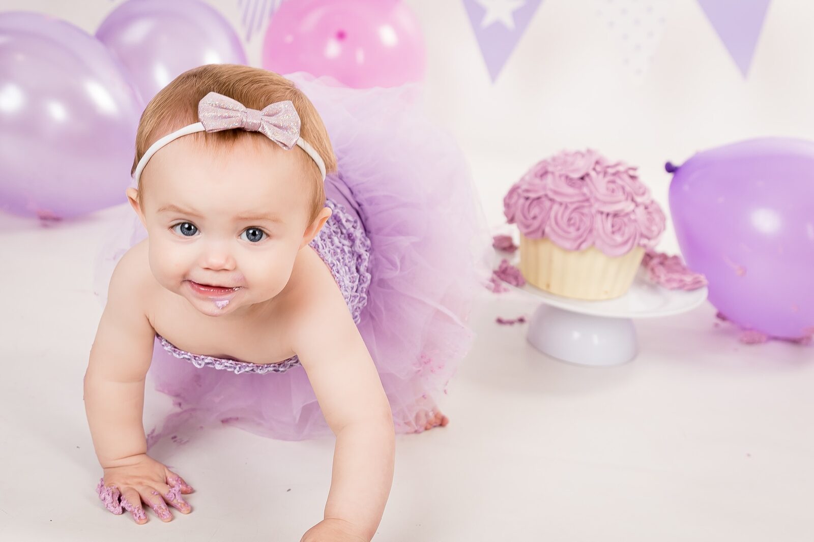 Smiling baby girl in lilac tutu and bow headband crawling beside a purple rosette cupcake with balloons and bunting