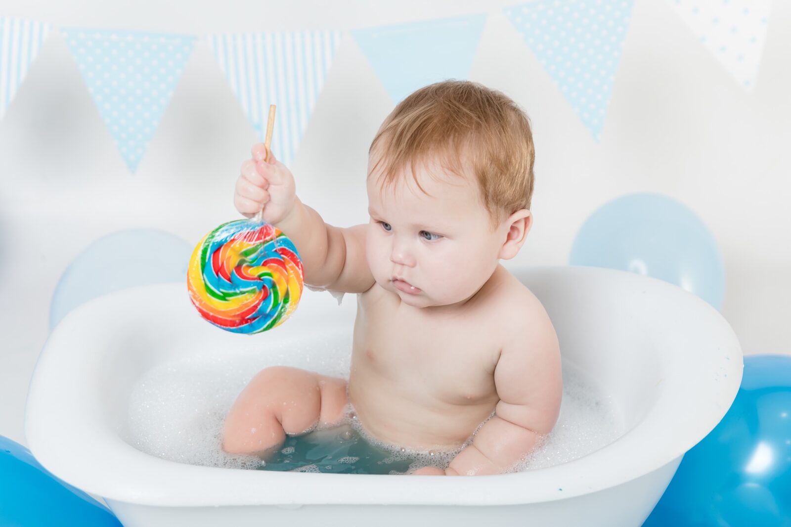 Baby sitting in a white bubble bath holding a colourful rainbow swirl lollipop with blue bunting and balloons