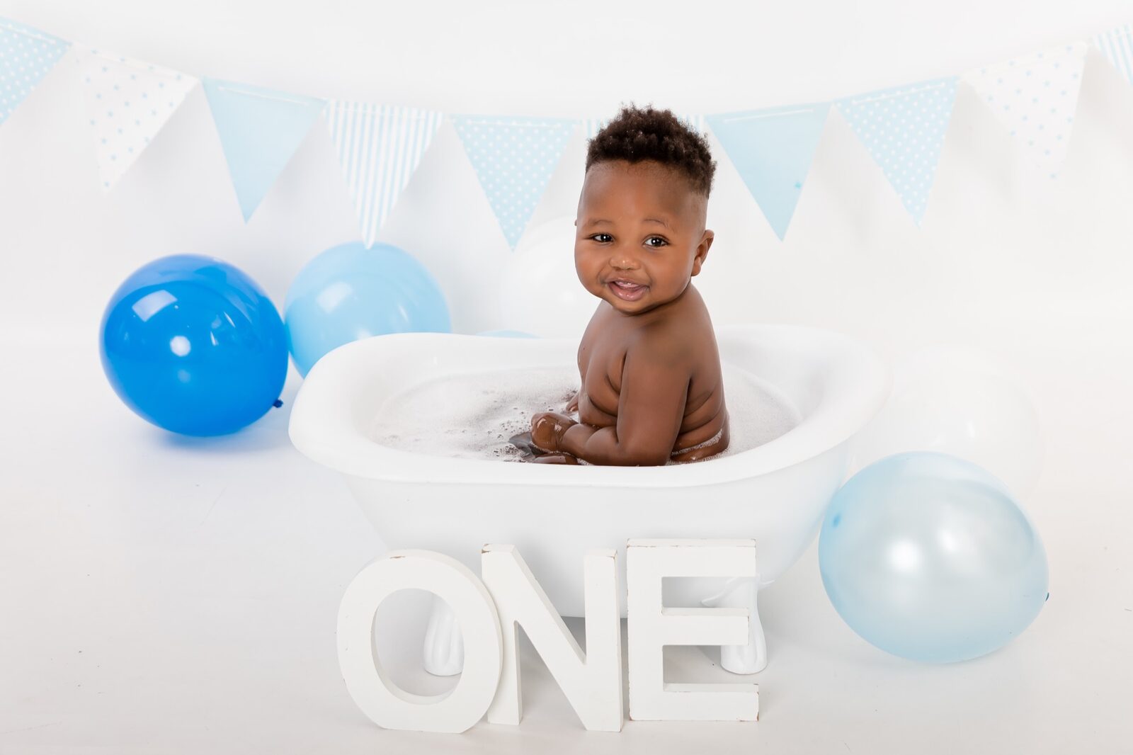 Smiling one-year-old baby boy sitting in a white bubble bath tub with blue balloons, bunting and ONE letters
