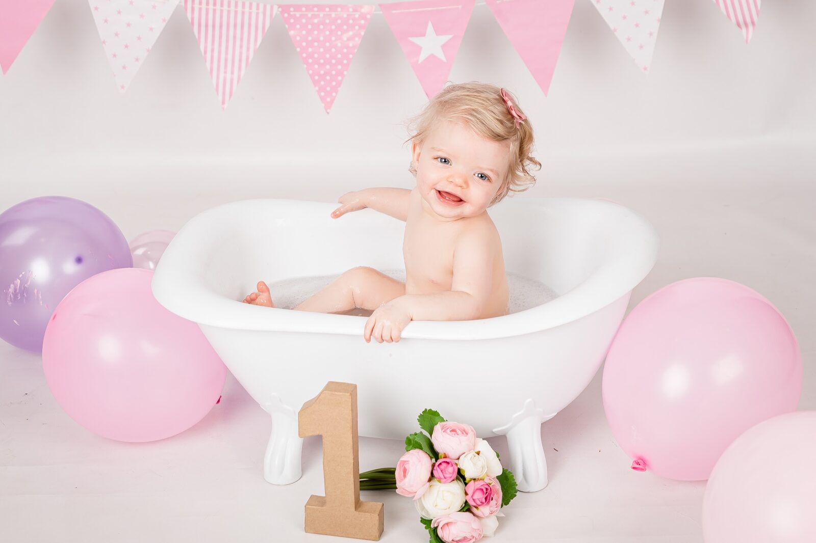 Blonde toddler girl smiling in a white clawfoot tub with pink bunting, balloons and a number one prop