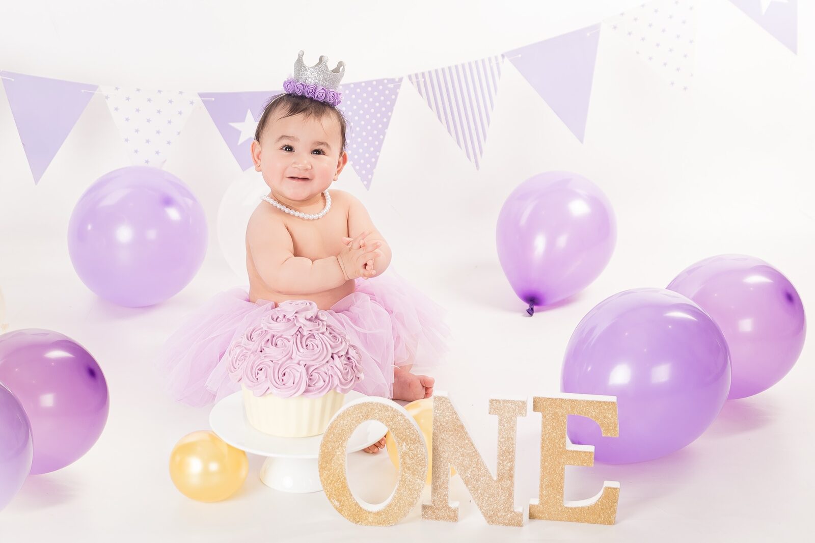 Smiling baby in pink tutu and silver crown sits beside a purple rosette cake with gold ONE letters and lilac balloons