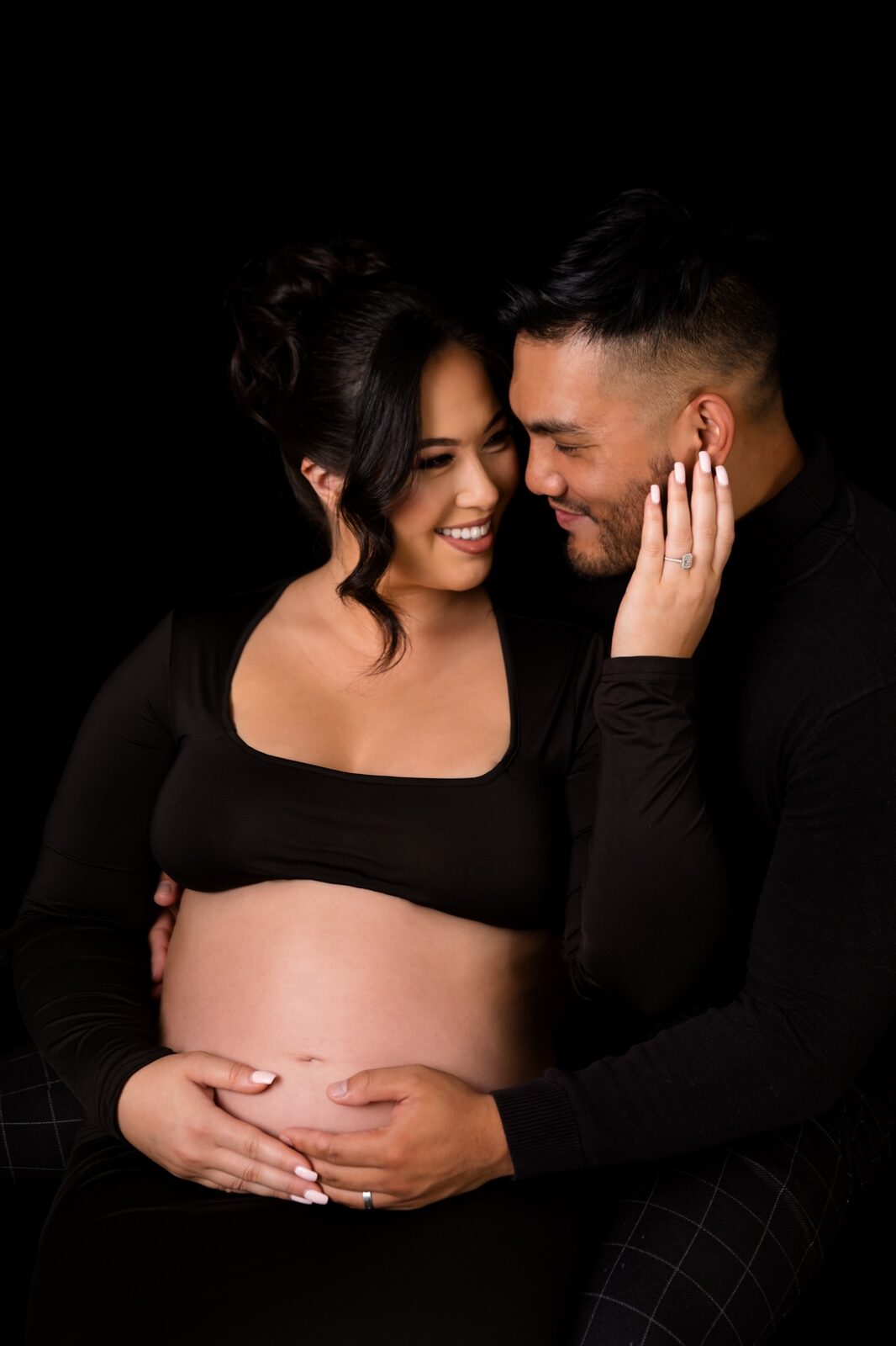 Pregnant woman in black crop top sharing a tender moment with her partner against a black studio backdrop