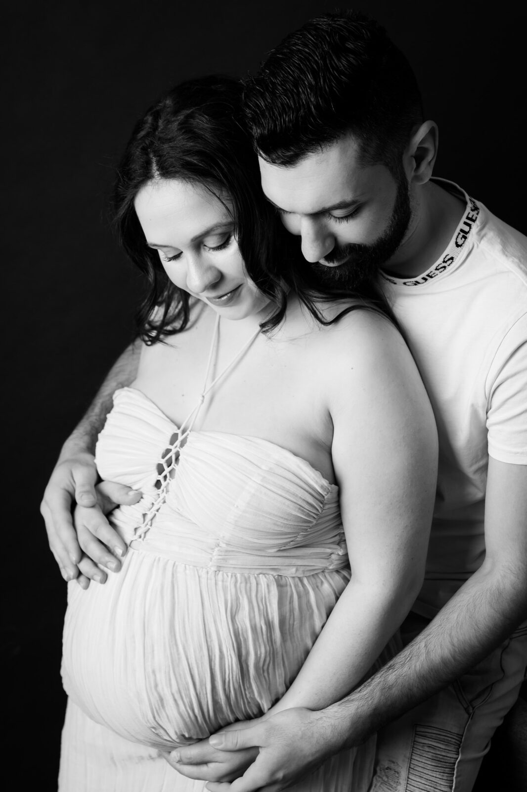 Black and white studio maternity photo of an expectant couple embracing, hands cradling the baby bump against a dark backdrop