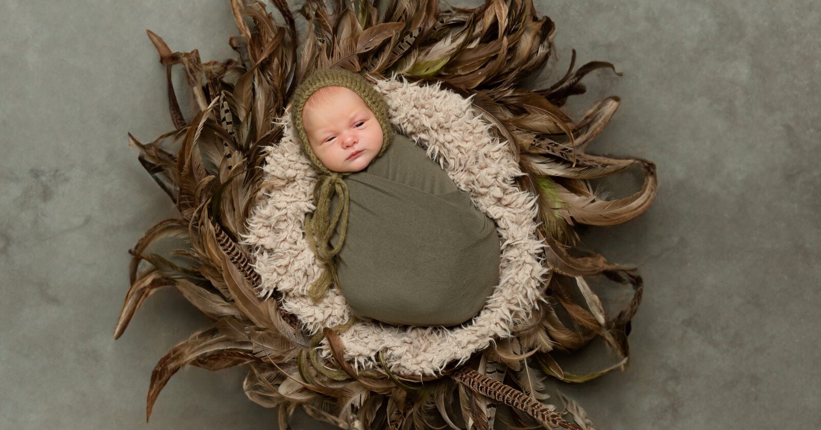 Newborn baby in olive green knit bonnet and swaddle, nestled in a cream fur-lined basket surrounded by brown pheasant feathe…
