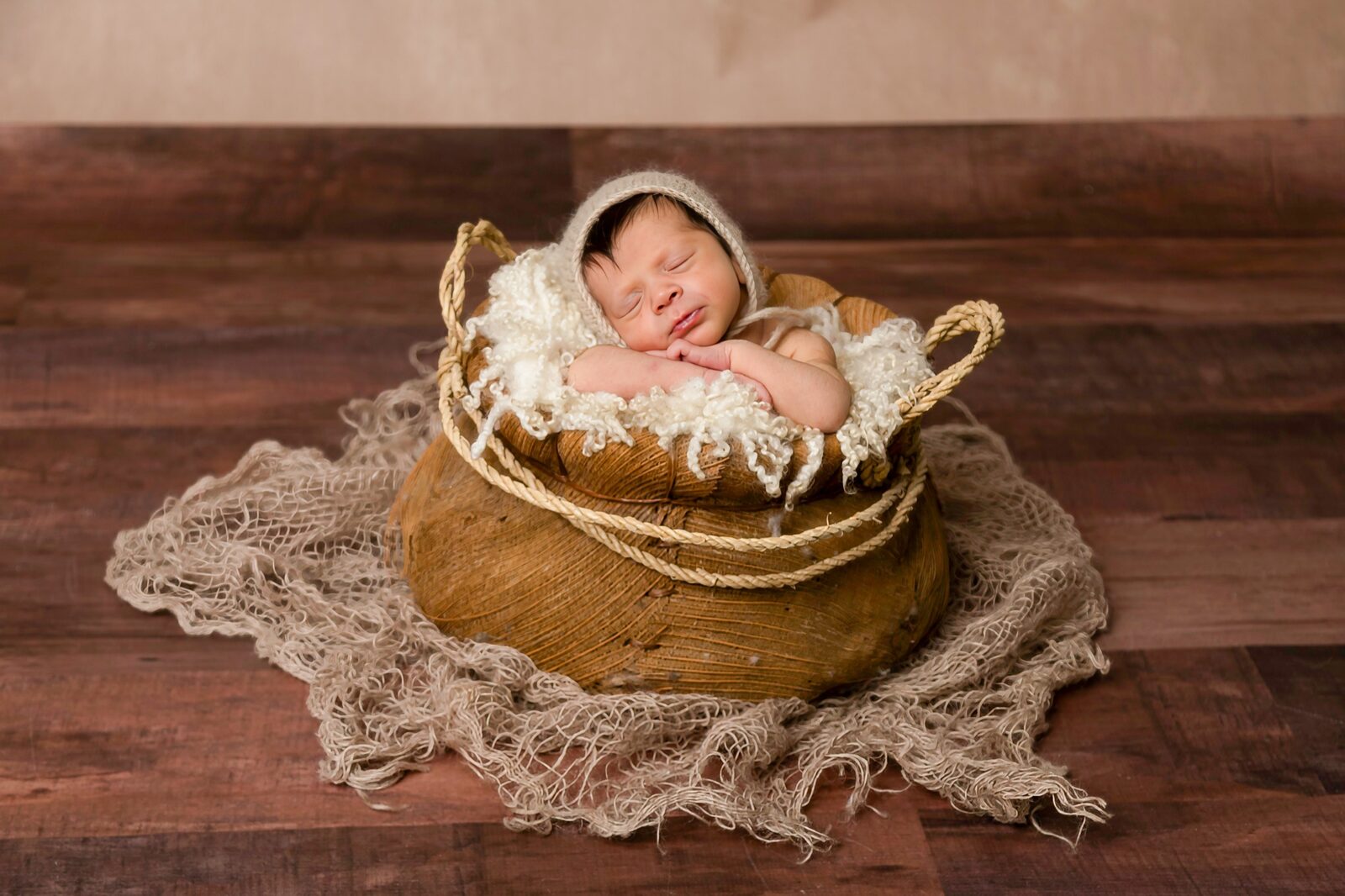 Newborn baby wearing a knitted bonnet sleeps peacefully in a rustic woven basket lined with cream wool on timber floor