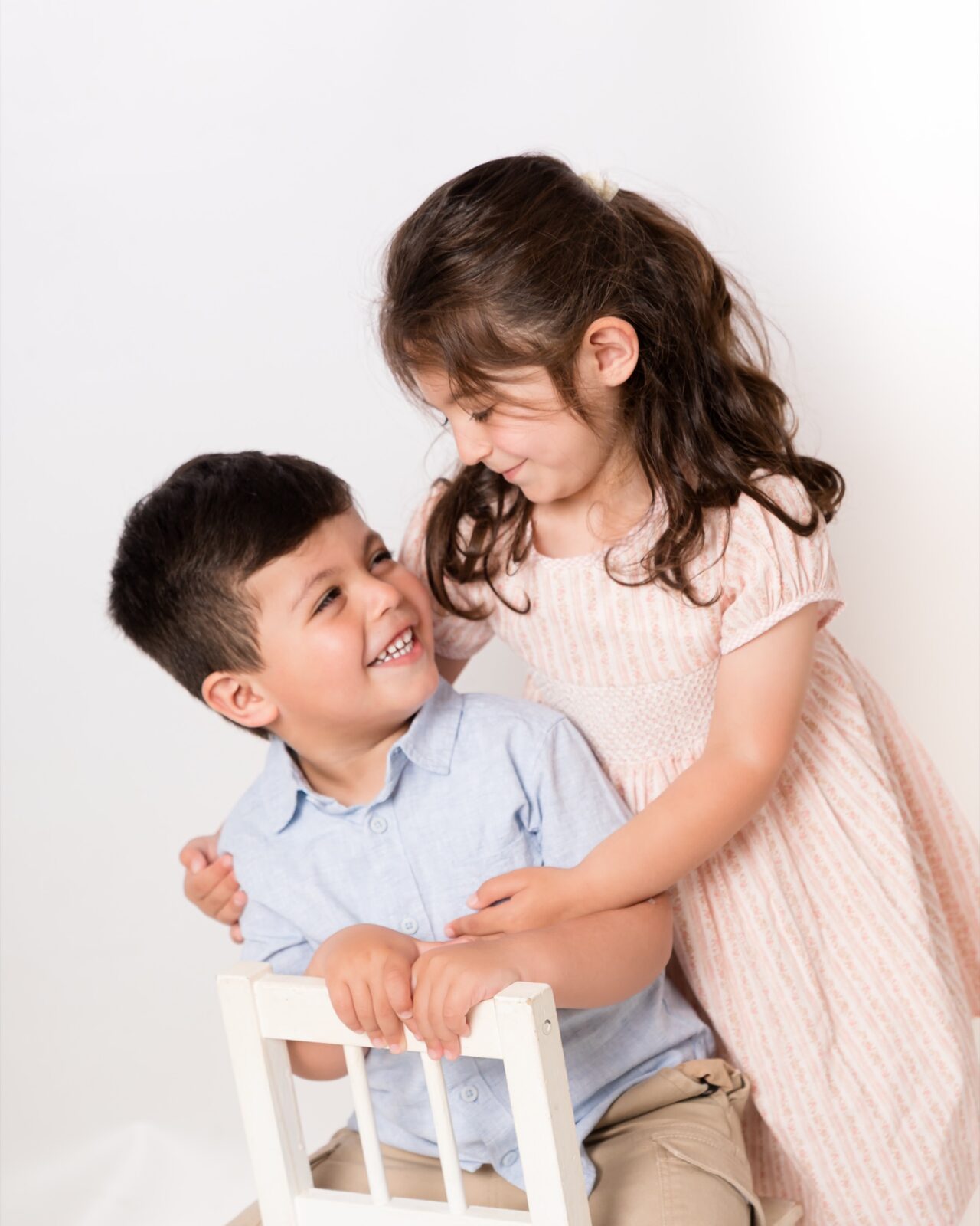 Young girl in pink dress hugging smiling boy in blue shirt seated on white chair in bright studio