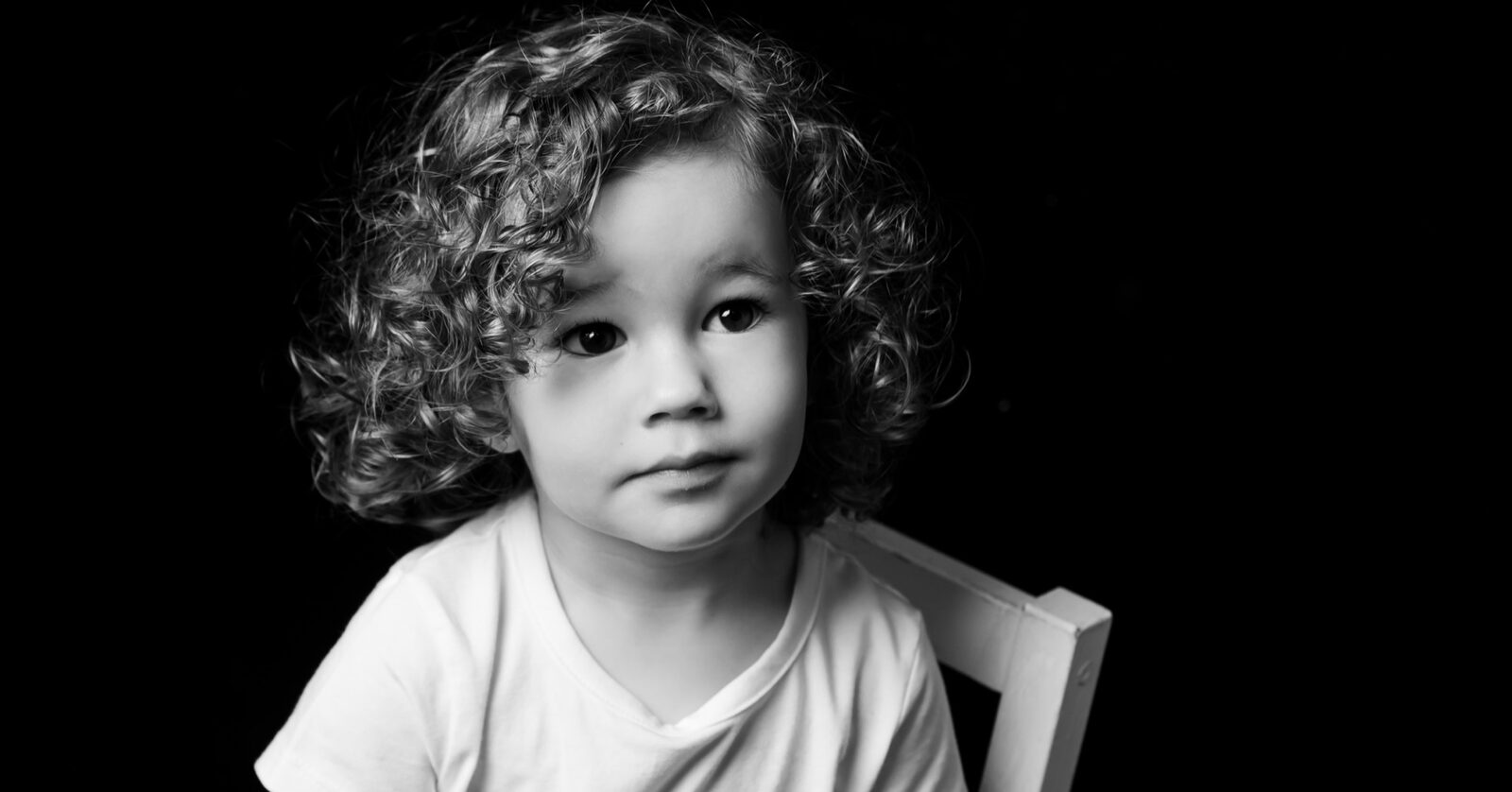 Monochrome studio portrait of a young child with curly hair, wearing a white t-shirt and seated on a wooden chair against a…