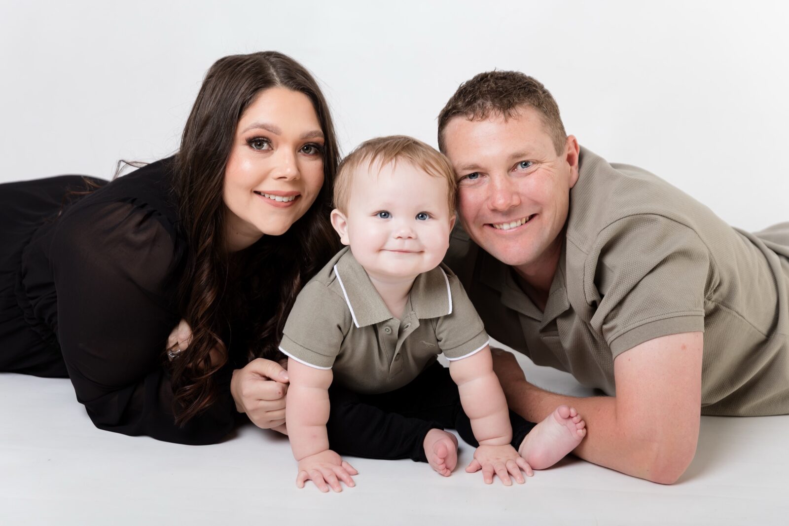 Mum, dad and baby boy lying on a white studio floor, all smiling at the camera in coordinated neutral outfits