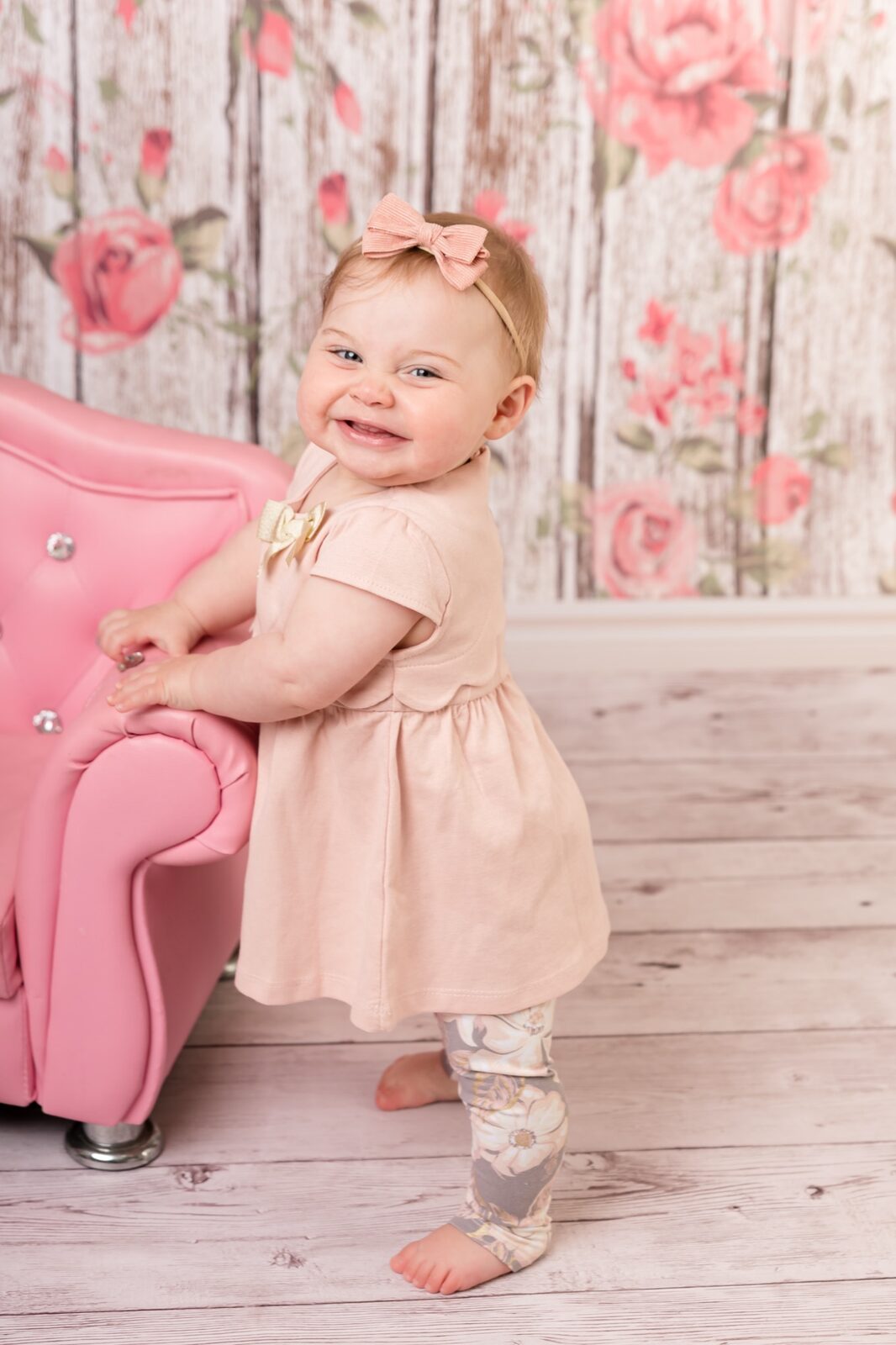 Baby girl in blush pink dress and floral leggings smiling beside a pink armchair in a floral studio set