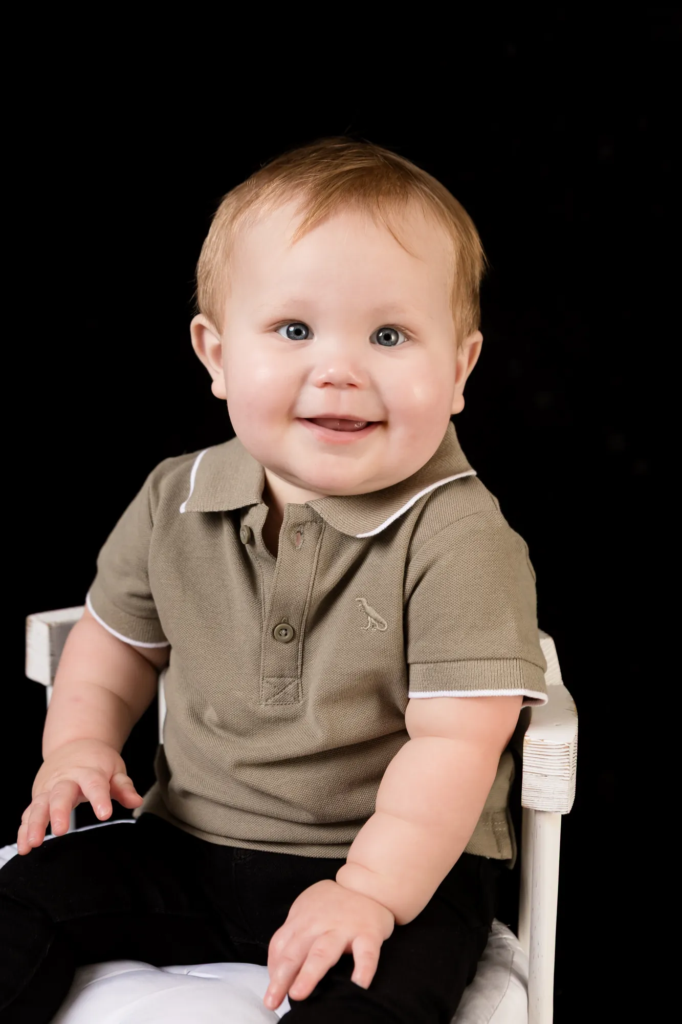 Smiling baby boy sitting up on a small white chair against a black studio backdrop at a sitter session