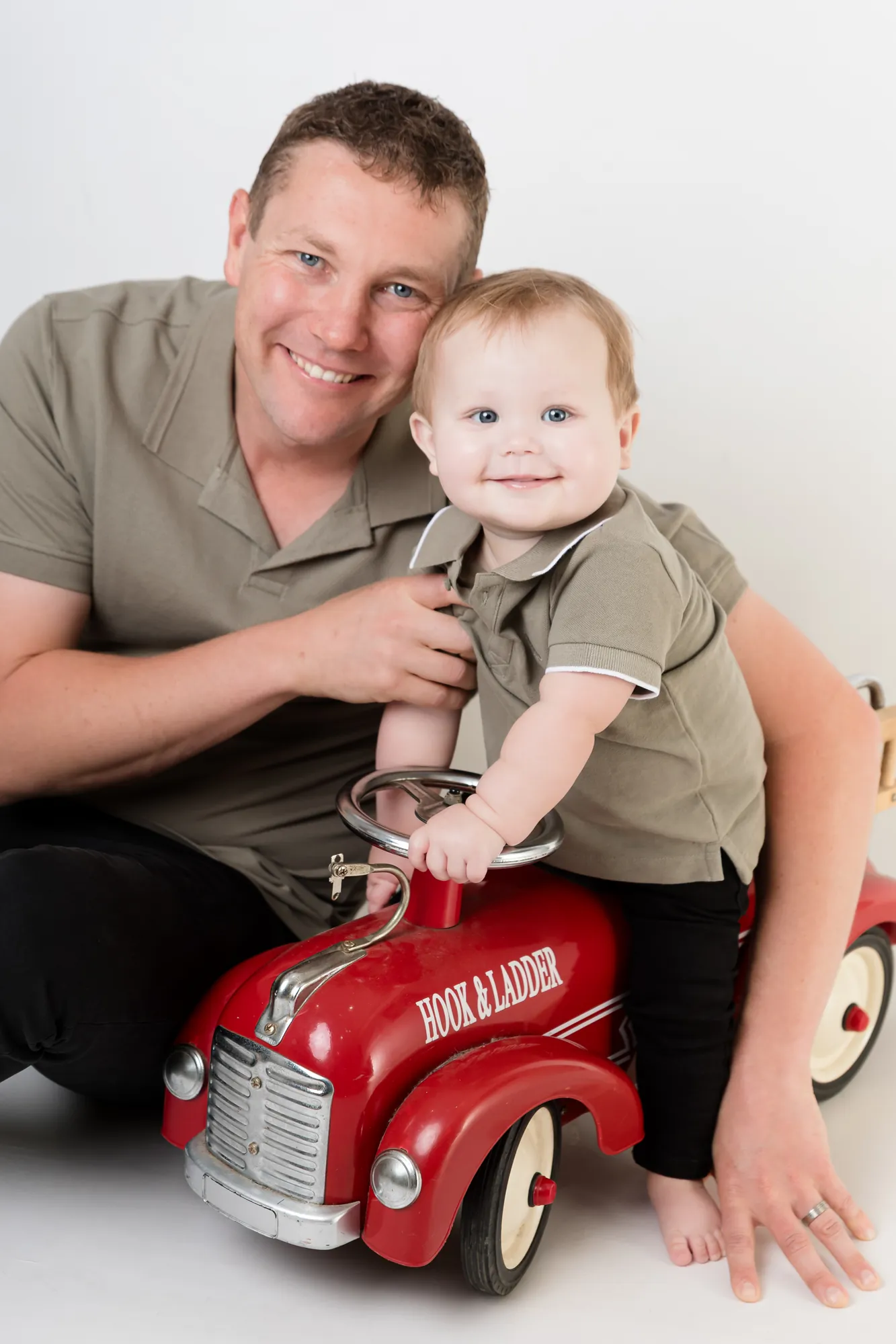 Dad crouching beside his smiling baby boy on a red ride-on truck