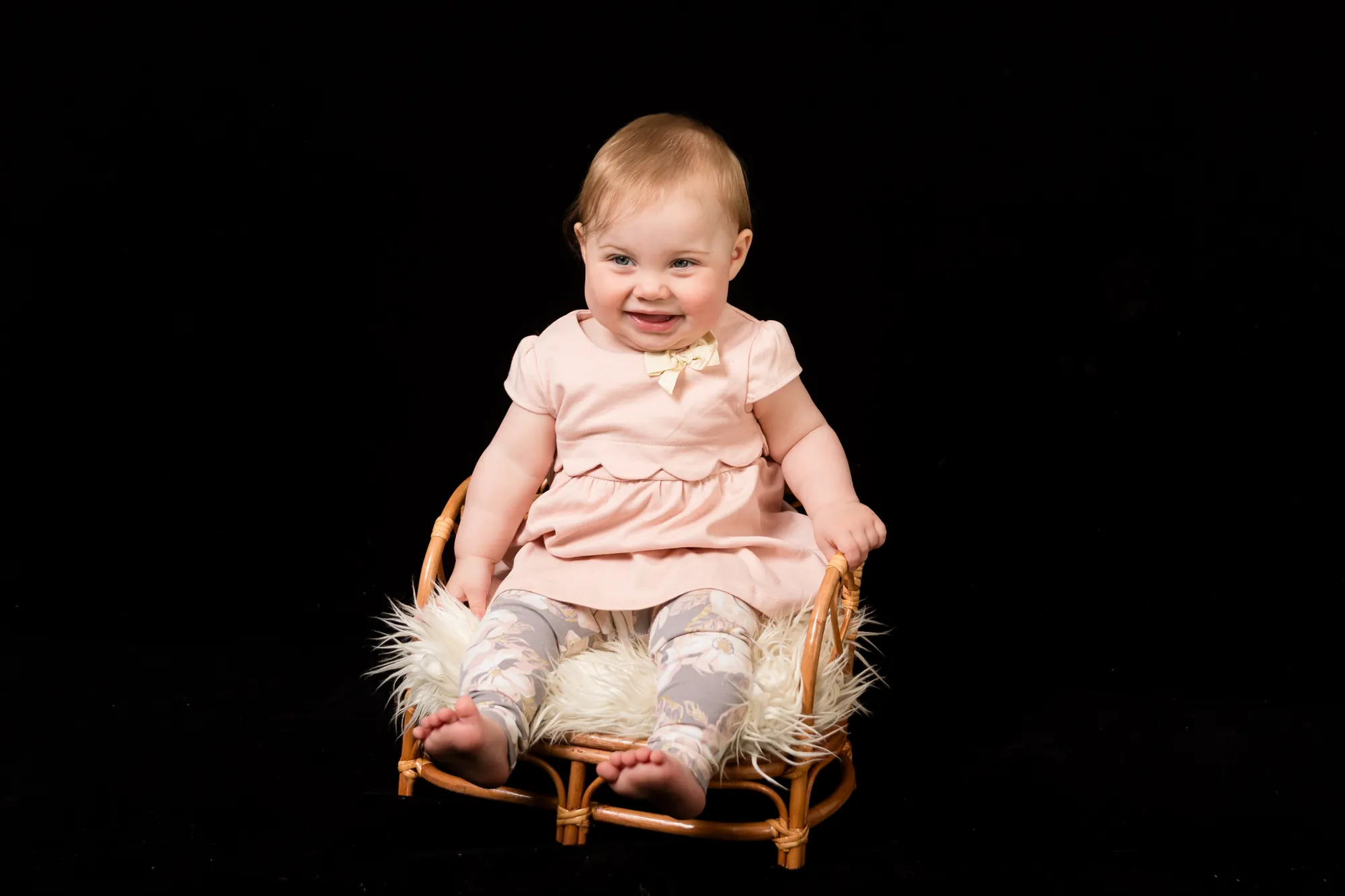 Baby girl in a pink dress sitting in a small wicker chair during a sitter photo session