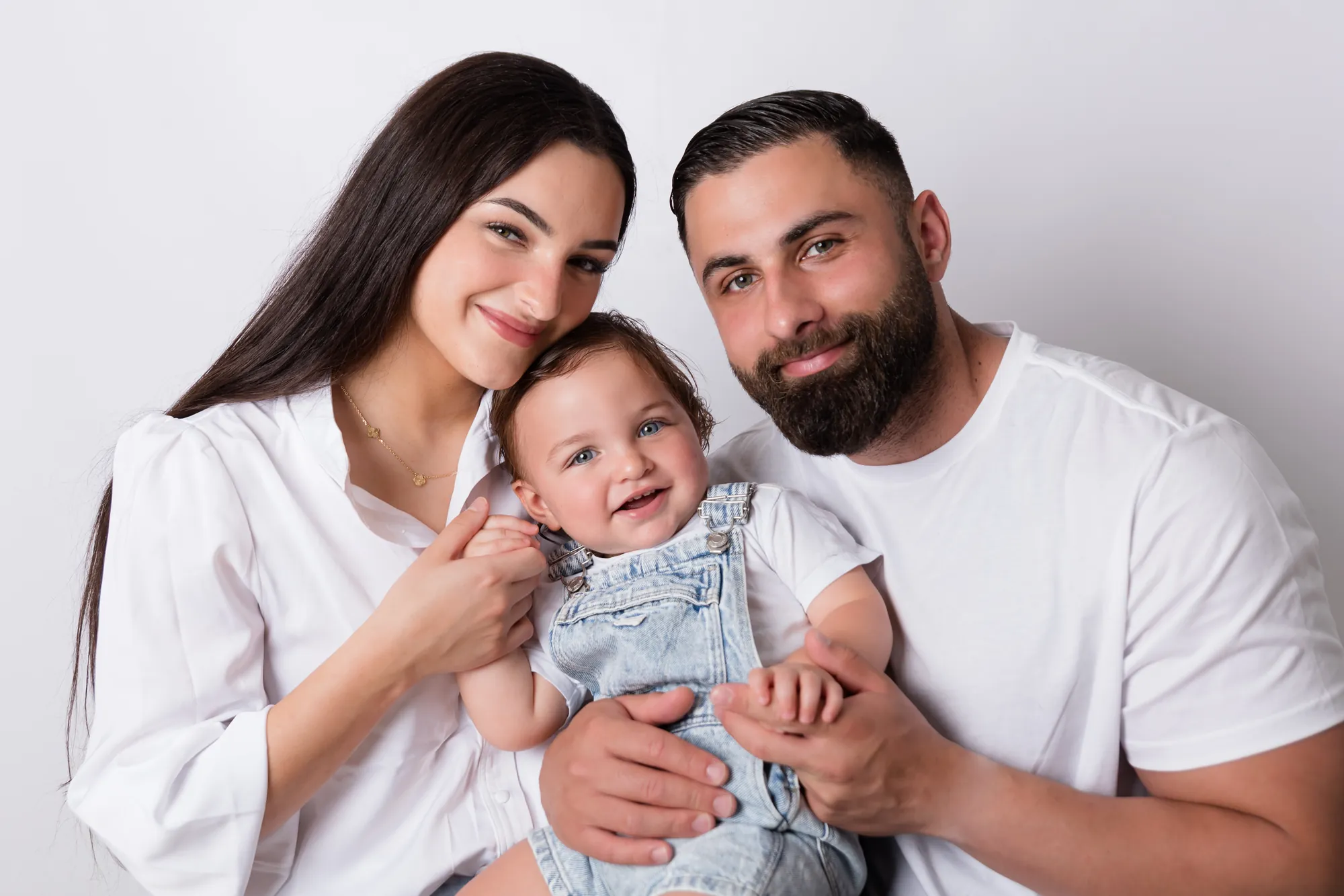 Mum and dad cuddling their smiling baby in denim overalls against a white backdrop