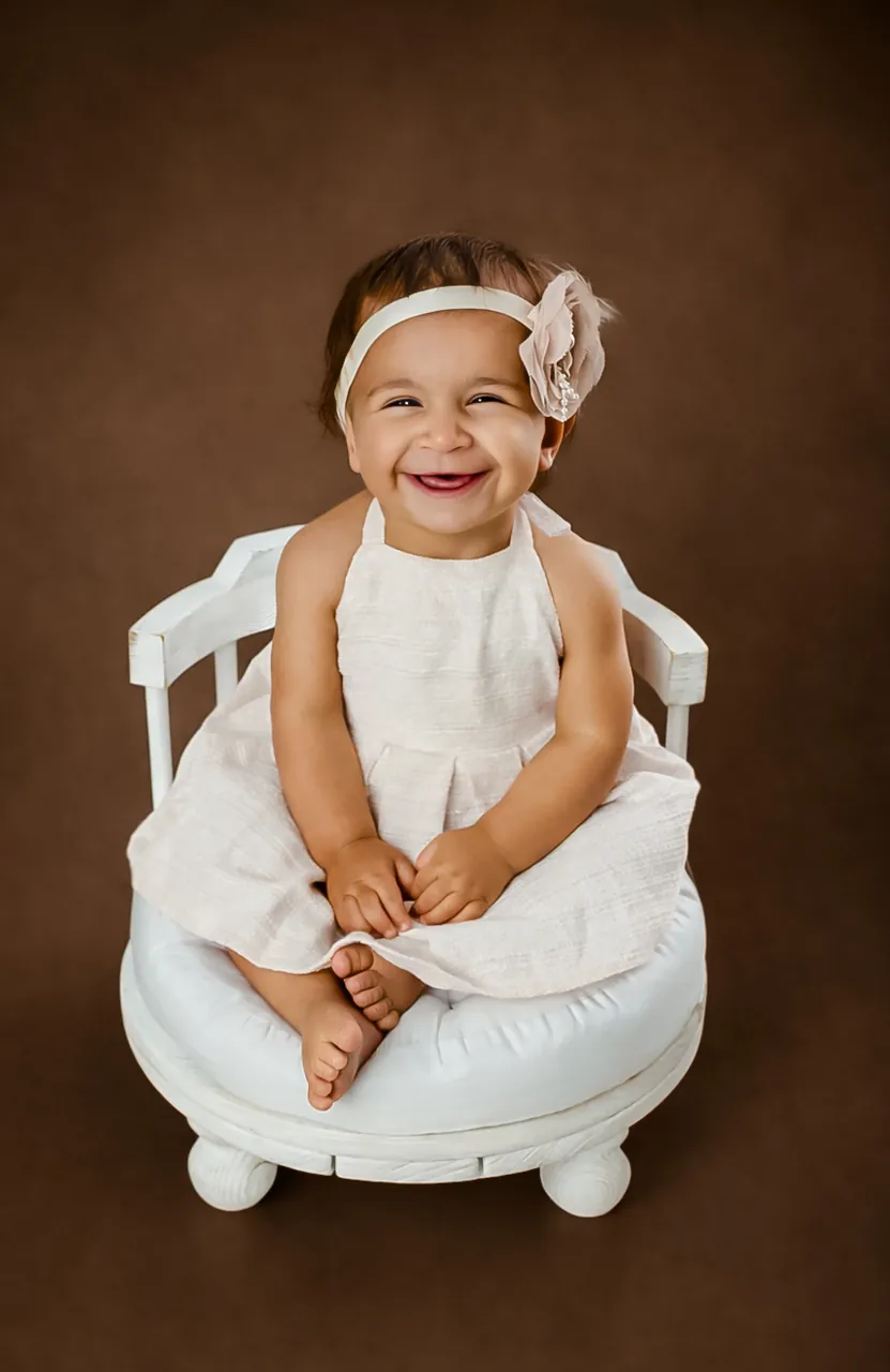 Laughing baby girl with a floral headband sitting on a small white seat against a brown backdrop